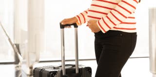 A pregnant person wearing a striped shirt is standing, holding their belly in one hand and the handle of a piece of luggage in the other hand