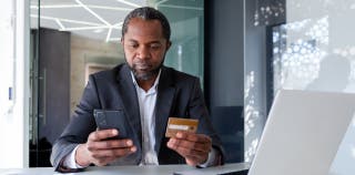 A man holds an American Express Business Gold Card in front of him with one hand and a smart phone with the other hand
