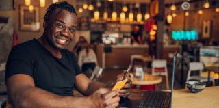 A smiling man sitting at a cafe table with an American Express Gold card in his hand and a laptop in front of him
