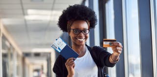 A woman smiling while holding a boarding pass in one hand and a credit card in the other, out in front of her