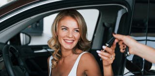A smiling blonde woman sitting inside of a car, being handed a set of keys