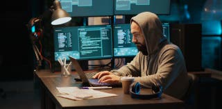 A hooded cyber hacker sits in front of multiple monitors, stealing data from a business