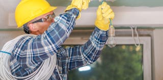 An electrician in a hard hat and protective gloves is installing a lighting fixture