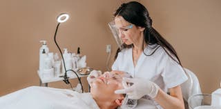 A female esthetician with a clear safety mask is giving a woman a facial