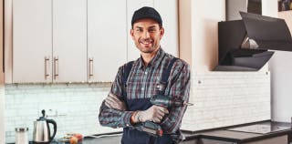 A handyman holding a drill stands with his arms crossed in front of a finished kitchen