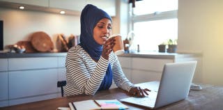 A woman in a hijab sits at a kitchen table with a cup of coffee in her hand and a laptop in front of her