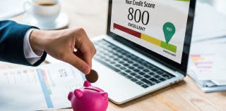 Hand putting coin in a piggy bank with a laptop in the background showing 800 credit score
