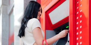 A brunette woman stands happily in front of a red ATM kiosk, taking out a cash advance with her credit card.