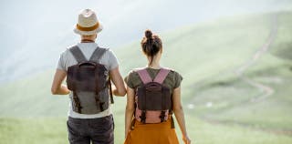 Two travelers with backpacks standing side by side, with their backs facing the camera. They are looking out at a mountain landscape.
