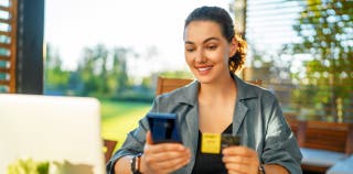 A woman smiling at her phone looking at her checking account and holding a debit card.