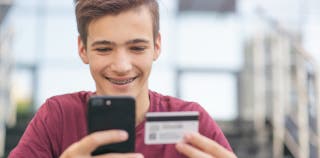 A teen smiling at their checking account on their phone holding a card in their hand. 