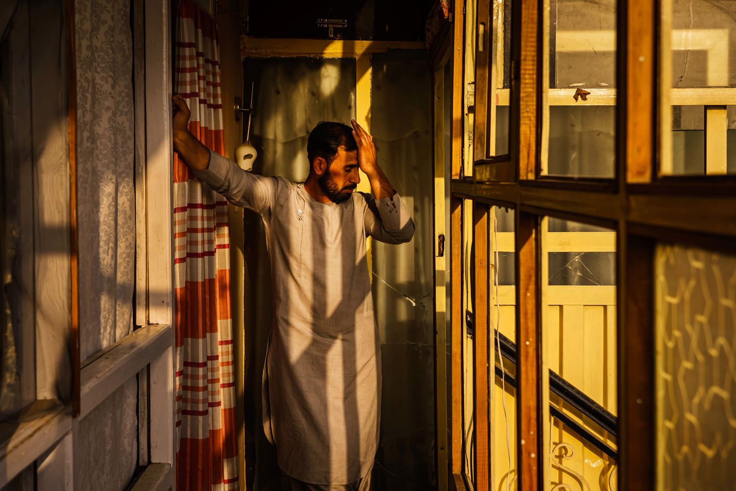 A man stands near a wall of windows with his hand held up to his forehead.
