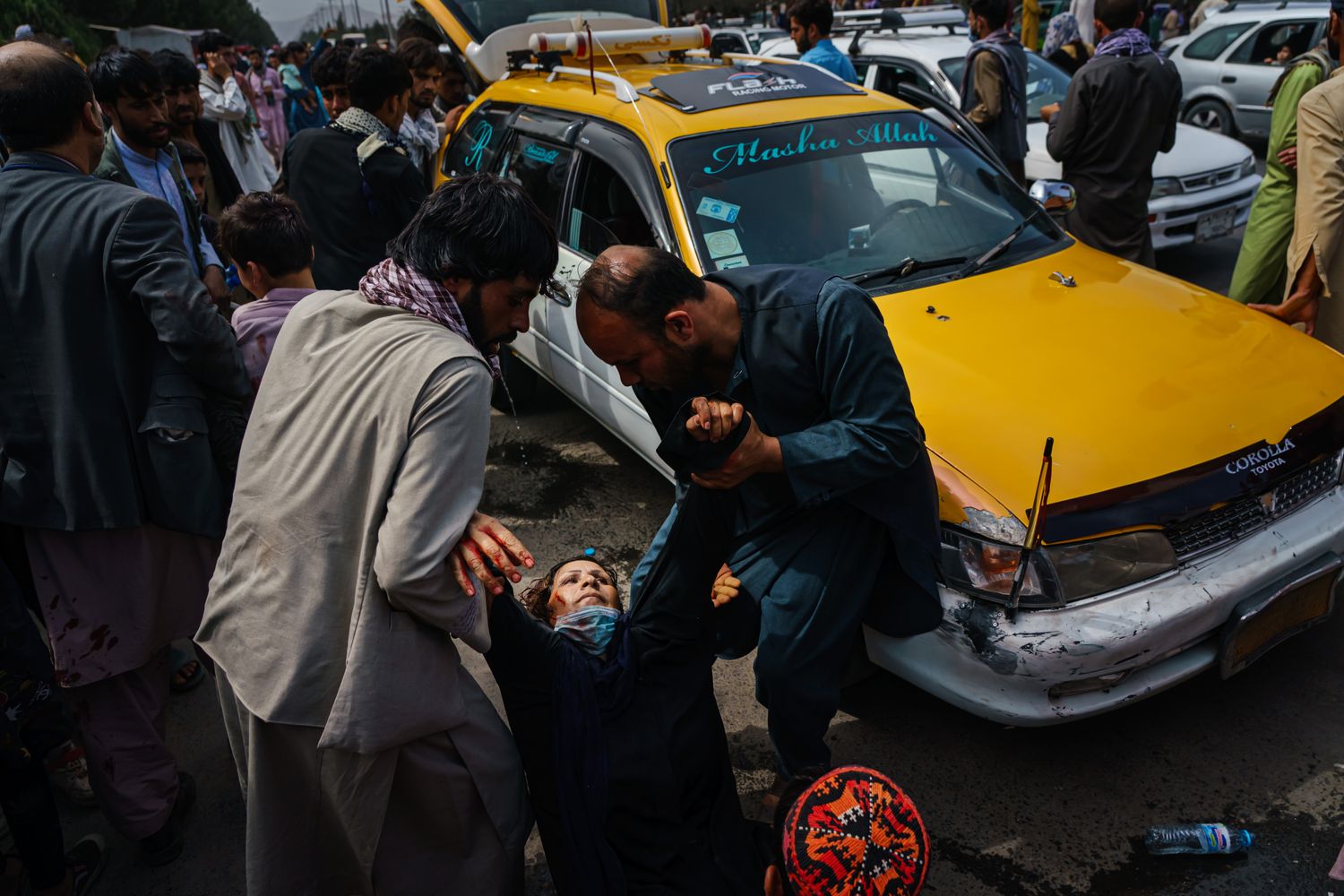 Two men pulling up a bloodied woman by her arms next to a yellow car.