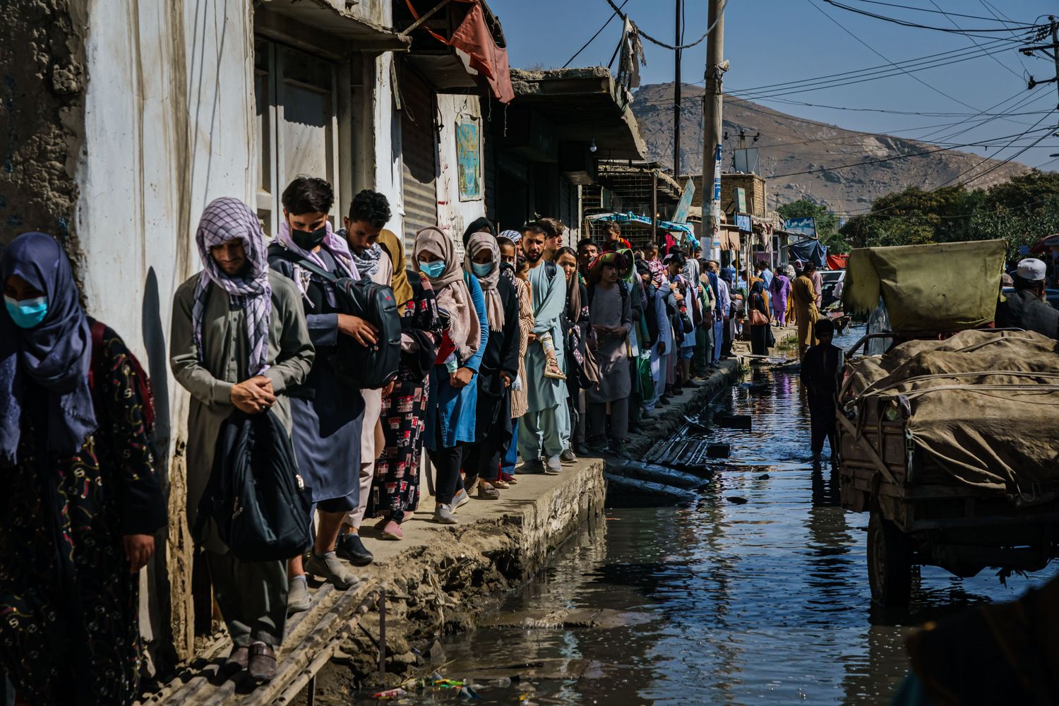 Afghans stand in a line along the edge of a flooded street.
