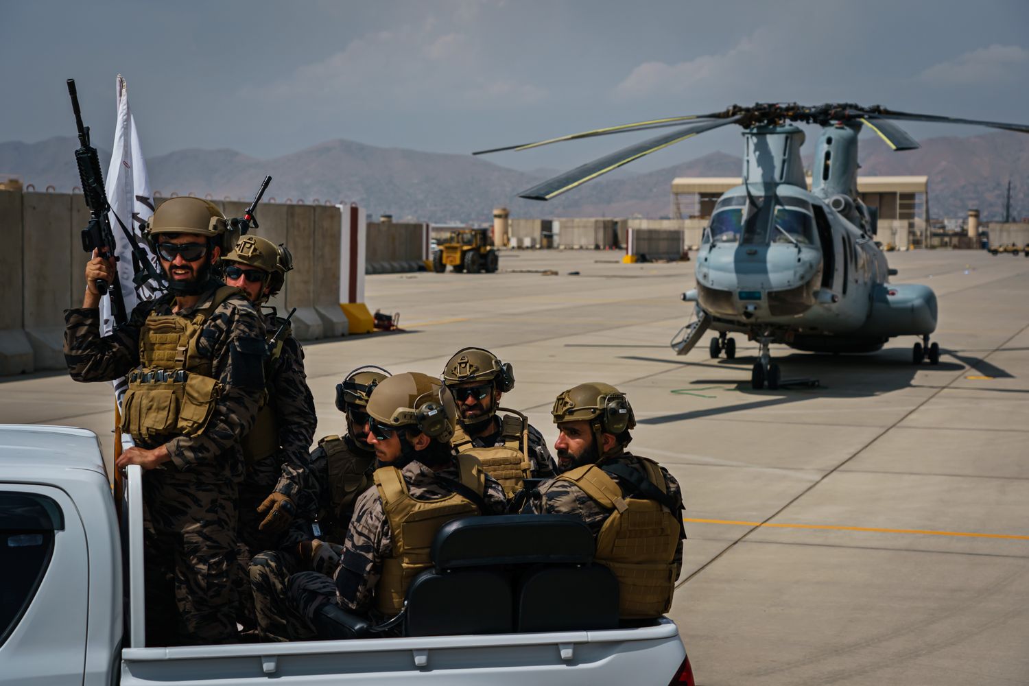 Taliban fighters sit in the back of a pickup truck next to an idle aircraft in daytime.