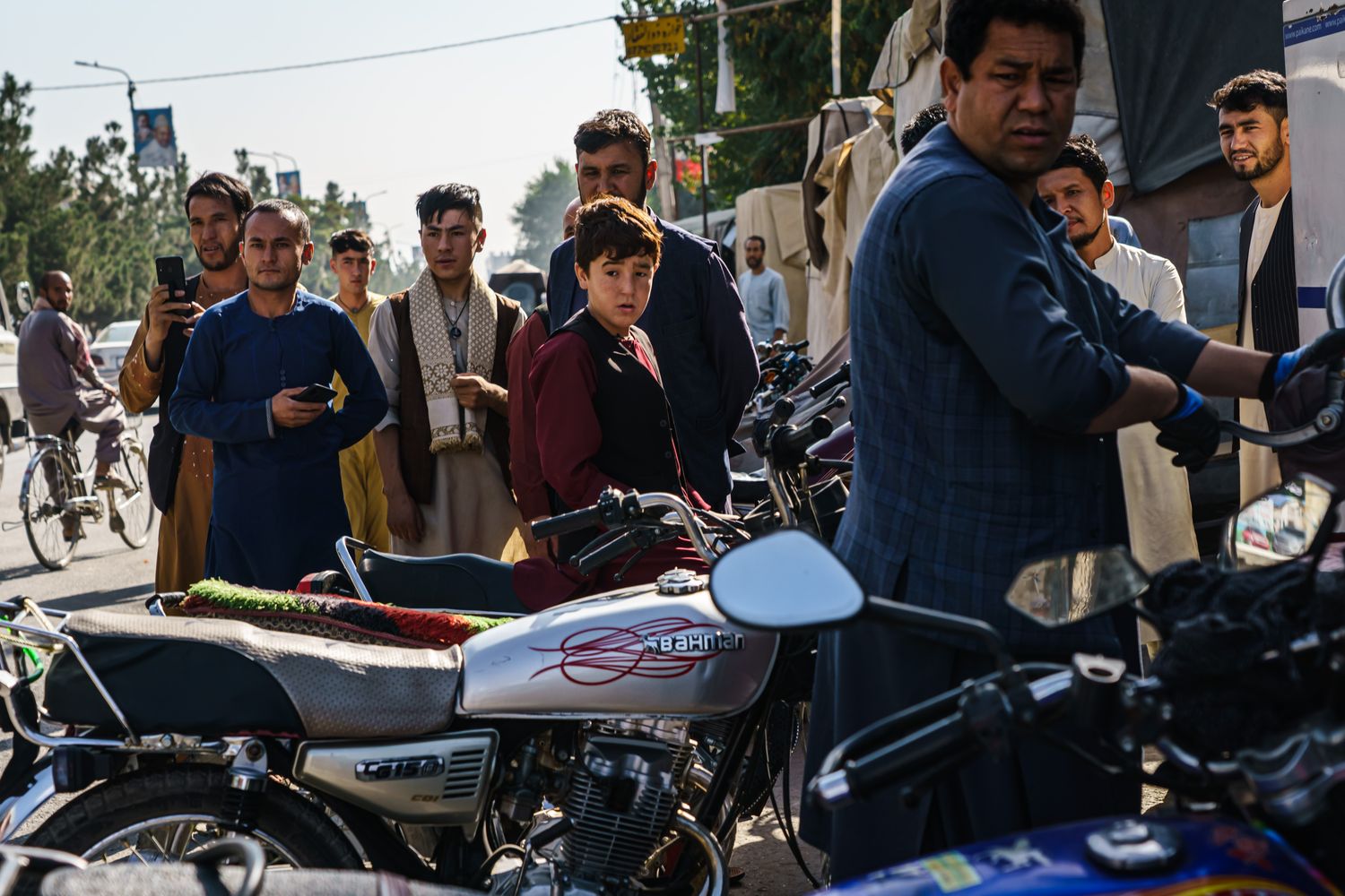 A group of men stand near motorcycles, one filming with his mobile phone.