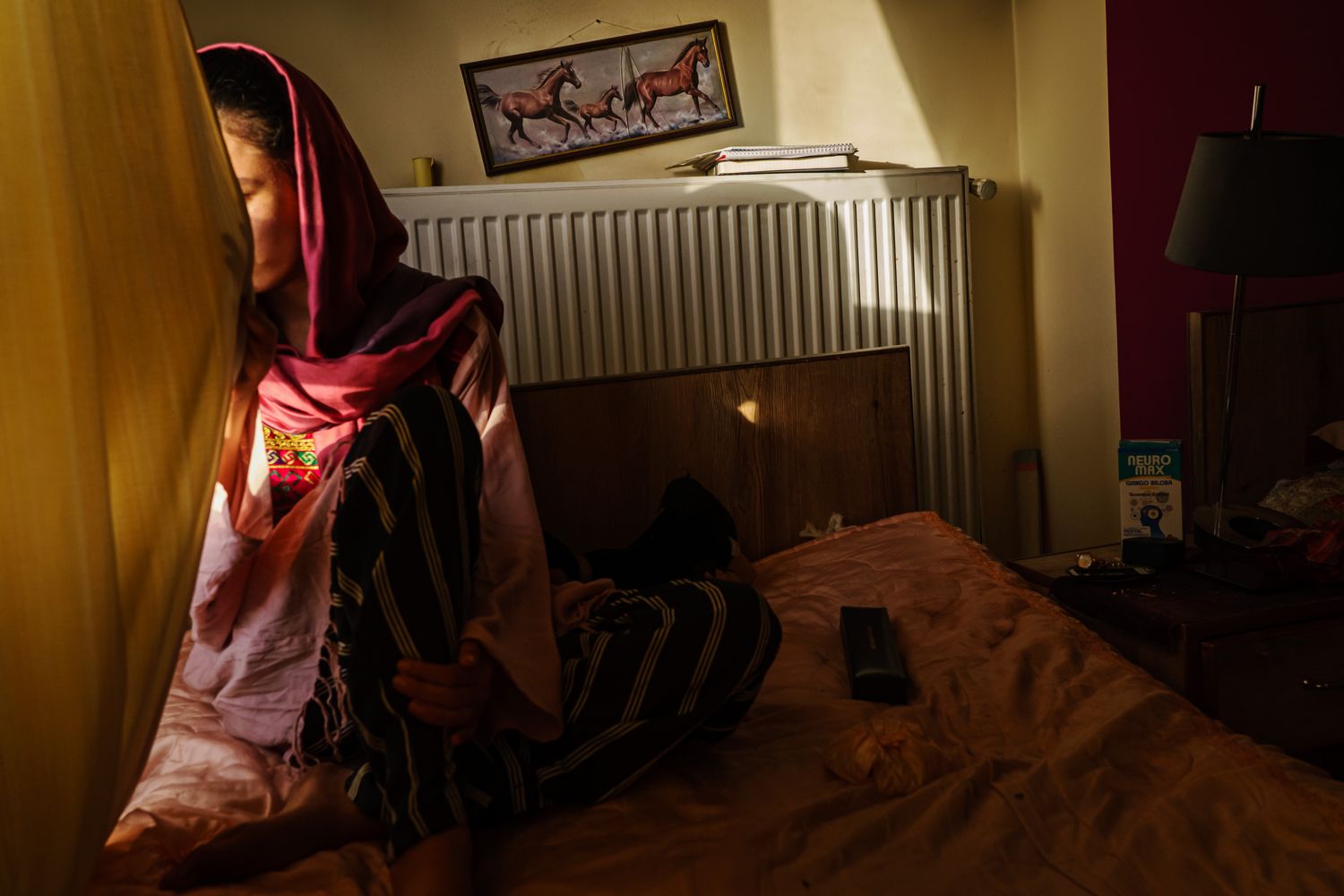A woman sits near a window with her face shielded by the drapes.