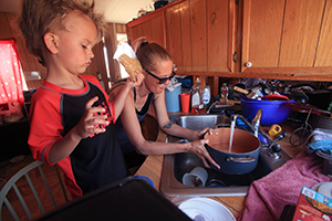 A young boy stands on a chair holding a bag of noodles while a woman in the background fills a pot with tap water.