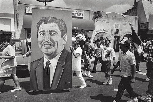 A group of people marching. Two people are carrying a large image of Ruben Salazar.