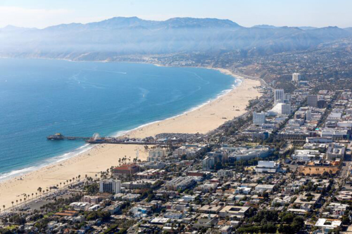 An aerial frame of Santa Monica, an urban area along a curved coast with a pier protruding into the ocean