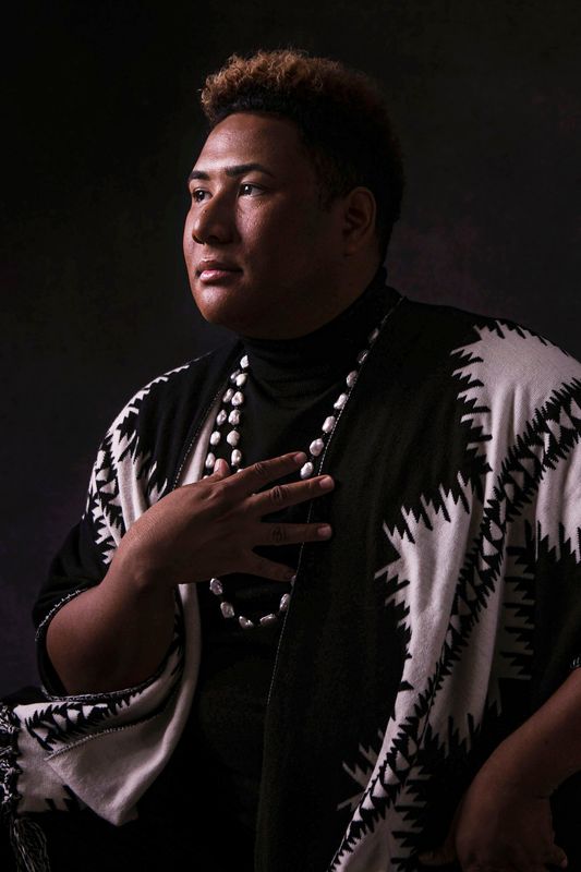Roger Q. Mason gestures to their chest, wearing a black and white shawl and necklace