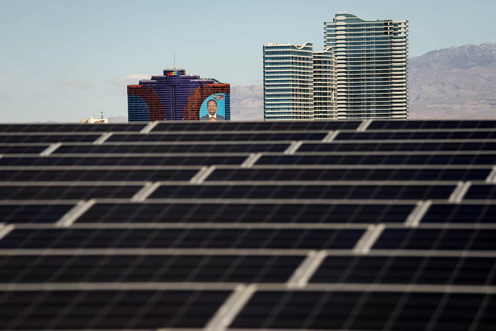 A long field of solar panels shimmer in front of a distant skyline of large buildings in the desert.