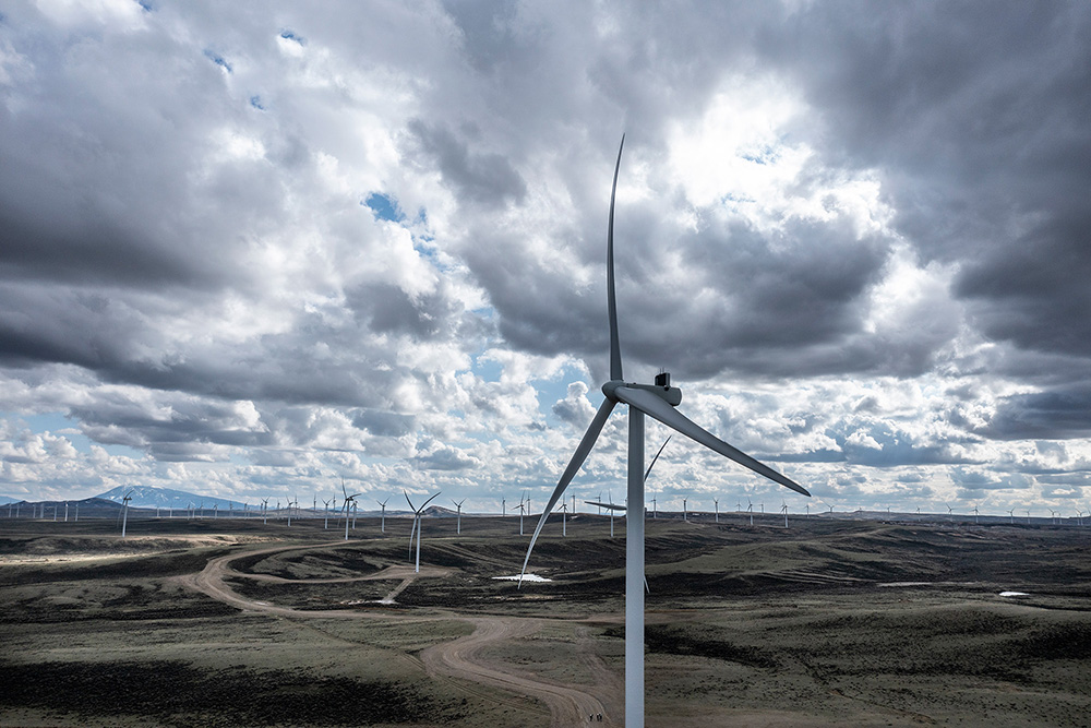 A landscape photo of a large wind turbine. In the backdrop a sprawling wind under a large cloudy sky
